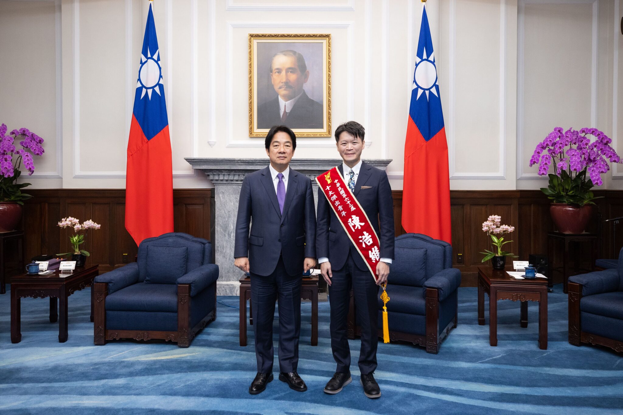 兩個西裝革履的男人站在一個房間里，房間里有旗幟和一張人的照片