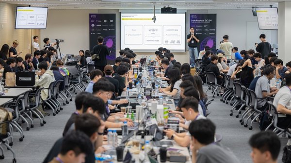 a group of people sitting at tables in a room with a screen and a person standing in front of them