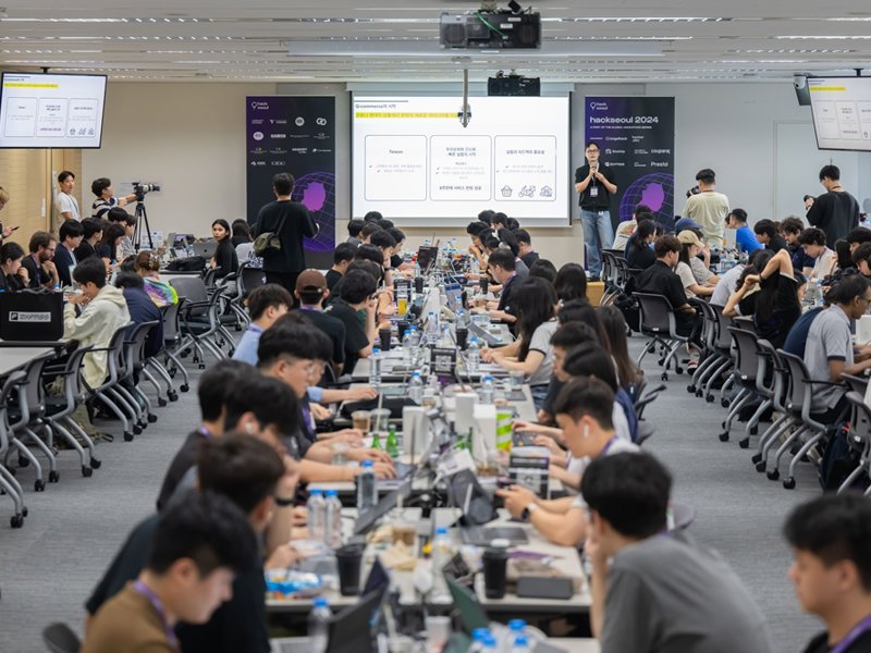 a group of people sitting at tables in a room with a screen and a person standing in front of them
