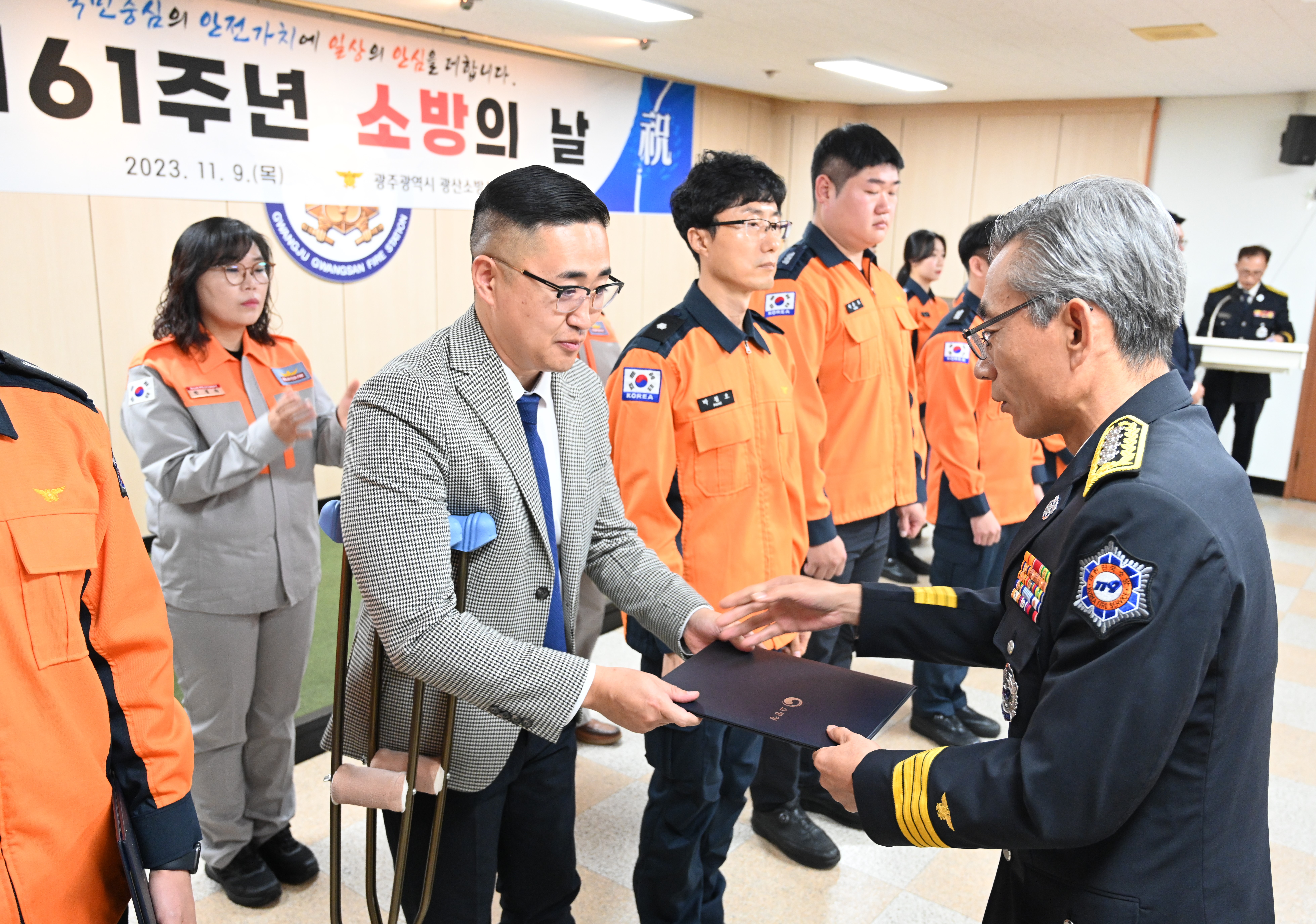 양복을 입은 사람이 다른 사람들과 함께 방에 있는 사람으로부터 증명서를 받고 있습니다.